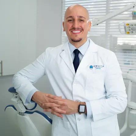 The image shows a smiling dental professional standing in front of a dental chair with a logo on his white coat that reads AMAYA, wearing glasses, and posing confidently for the camera.