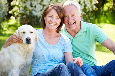 A man and woman are sitting on grass with a golden retriever between them they all appear happy and relaxed, likely enjoying a sunny day outdoors.